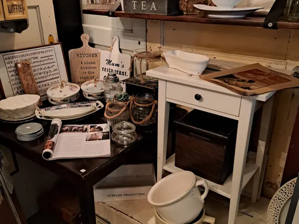 Kringloopwinkel interieur met huishoudartikelen: zwarte tafel met servies, witte fornuis, planken met decoratieve items en ke