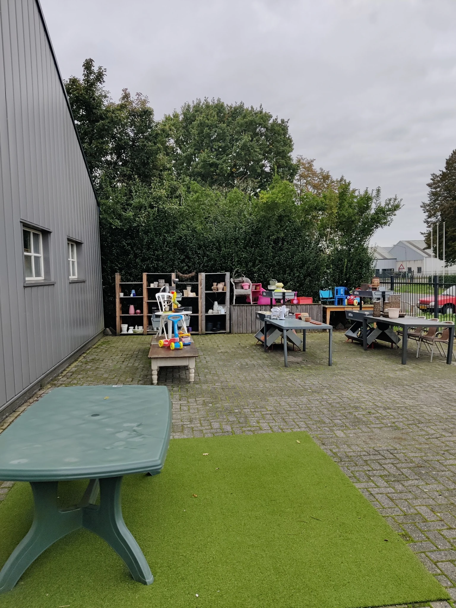 Buitenterrein met kasten, tafels, speelgoed en diverse kringloopwaar langs een grijze loods