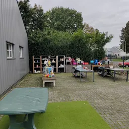 Buitenterrein met kasten, tafels, speelgoed en diverse kringloopwaar langs een grijze loods