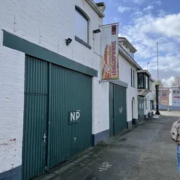 Witte bakstenen gevel van kringloopwinkel met groene metalen deuren en 'NP' markering, aan een straat