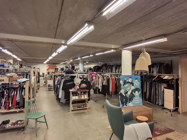 Ruim interieur van kringloopwinkel met kledingrekken, zithoek met groene stoel, hangende lamp en betonnen plafond