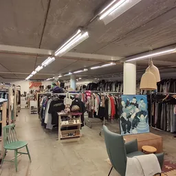 Ruim interieur van kringloopwinkel met kledingrekken, zithoek met groene stoel, hangende lamp en betonnen plafond