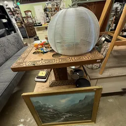 Verschillende meubels en huisraad uitgestald in de winkel, waaronder een tafel met inlegwerk, een witte hanglamp en een schil