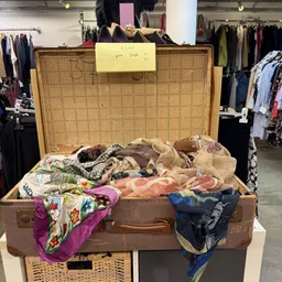 Geopende bruine vintage koffer vol met tweedehands kleding en textiel, met geel prijskaartje eraan.