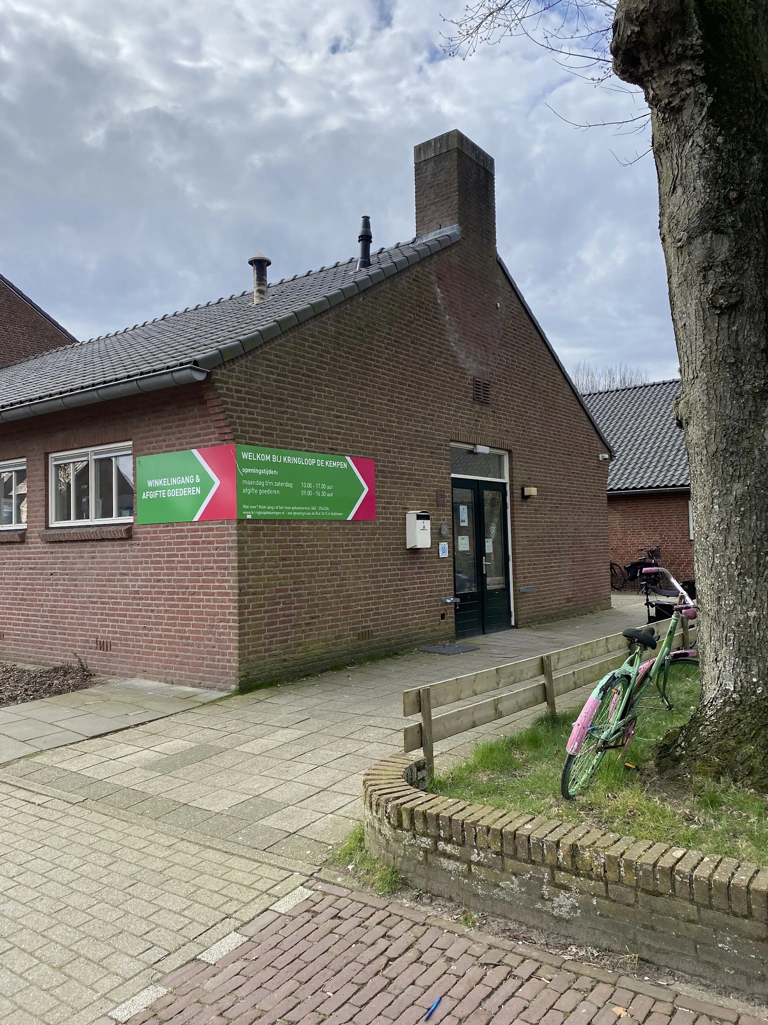 Bakstenen gevel van kringloopwinkel met groene en rode bewegwijzering borden, ingang met zwarte deur, fiets tegen boom
