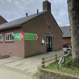 Bakstenen gevel van kringloopwinkel met groene en rode bewegwijzering borden, ingang met zwarte deur, fiets tegen boom