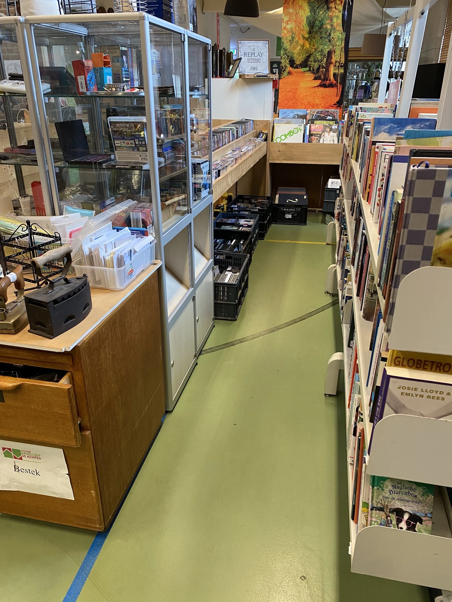 Winkelgangetje met stellingen vol boeken en media aan beide zijden, groene vloer, zwarte kratten in het midden