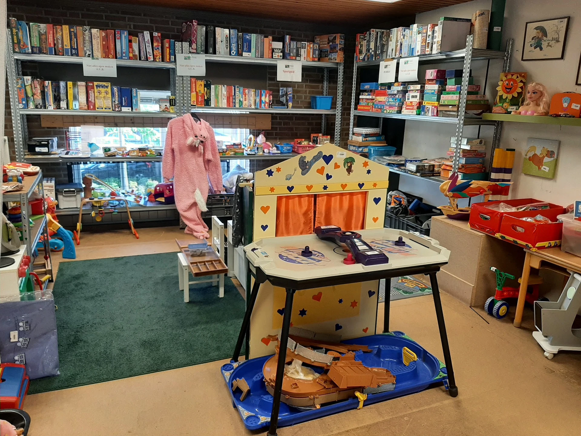 Kinderspeelhoek in kringloopwinkel met speelgoed, boeken in stellingen op achtergrond en groene vloerbedekking