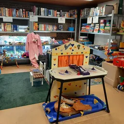 Kinderspeelhoek in kringloopwinkel met speelgoed, boeken in stellingen op achtergrond en groene vloerbedekking
