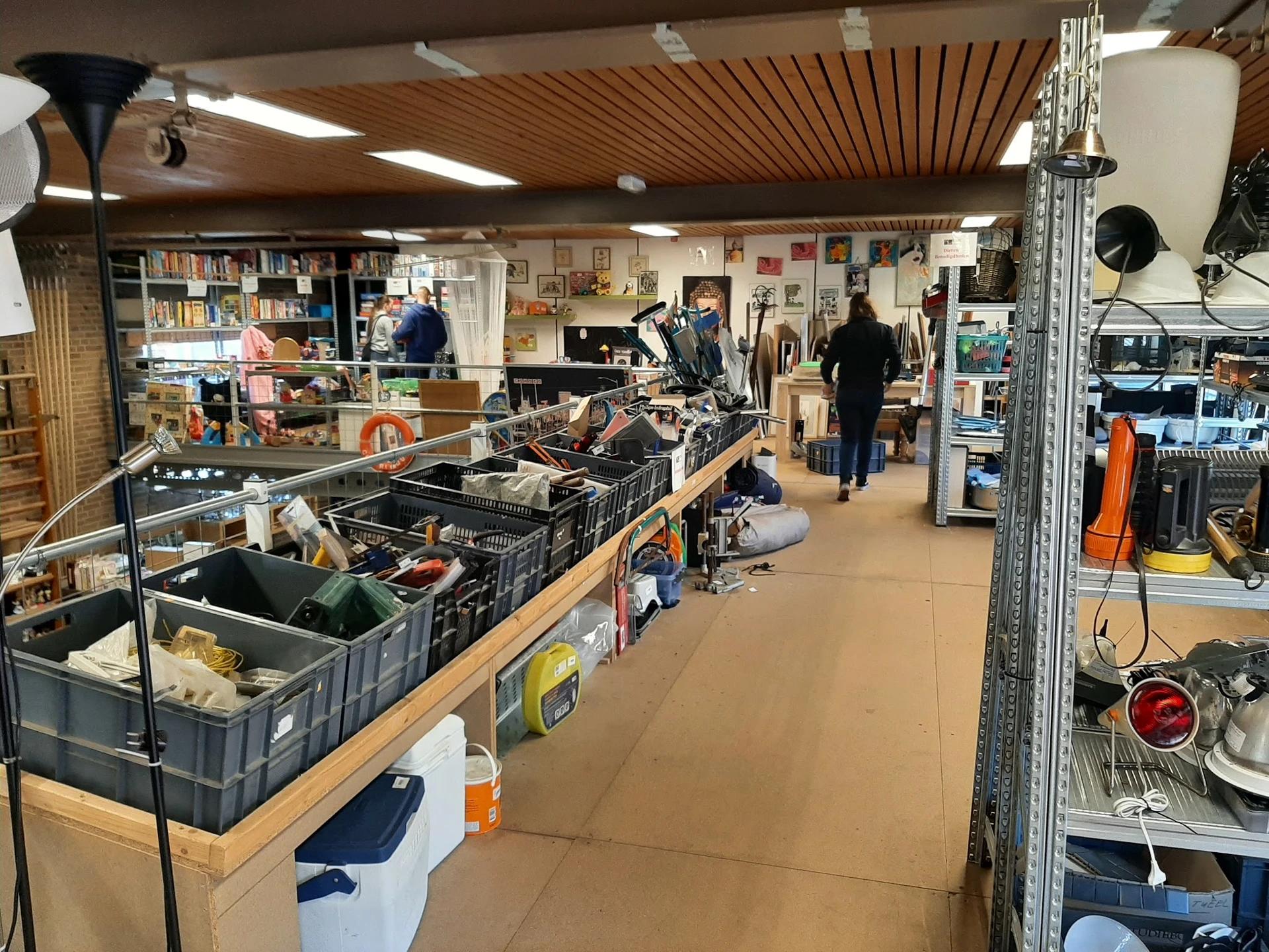 Overzicht van de winkelvloer met stellingen, zwarte kratten met spullen, kasten met artikelen en bezoekers in de kringloopwin