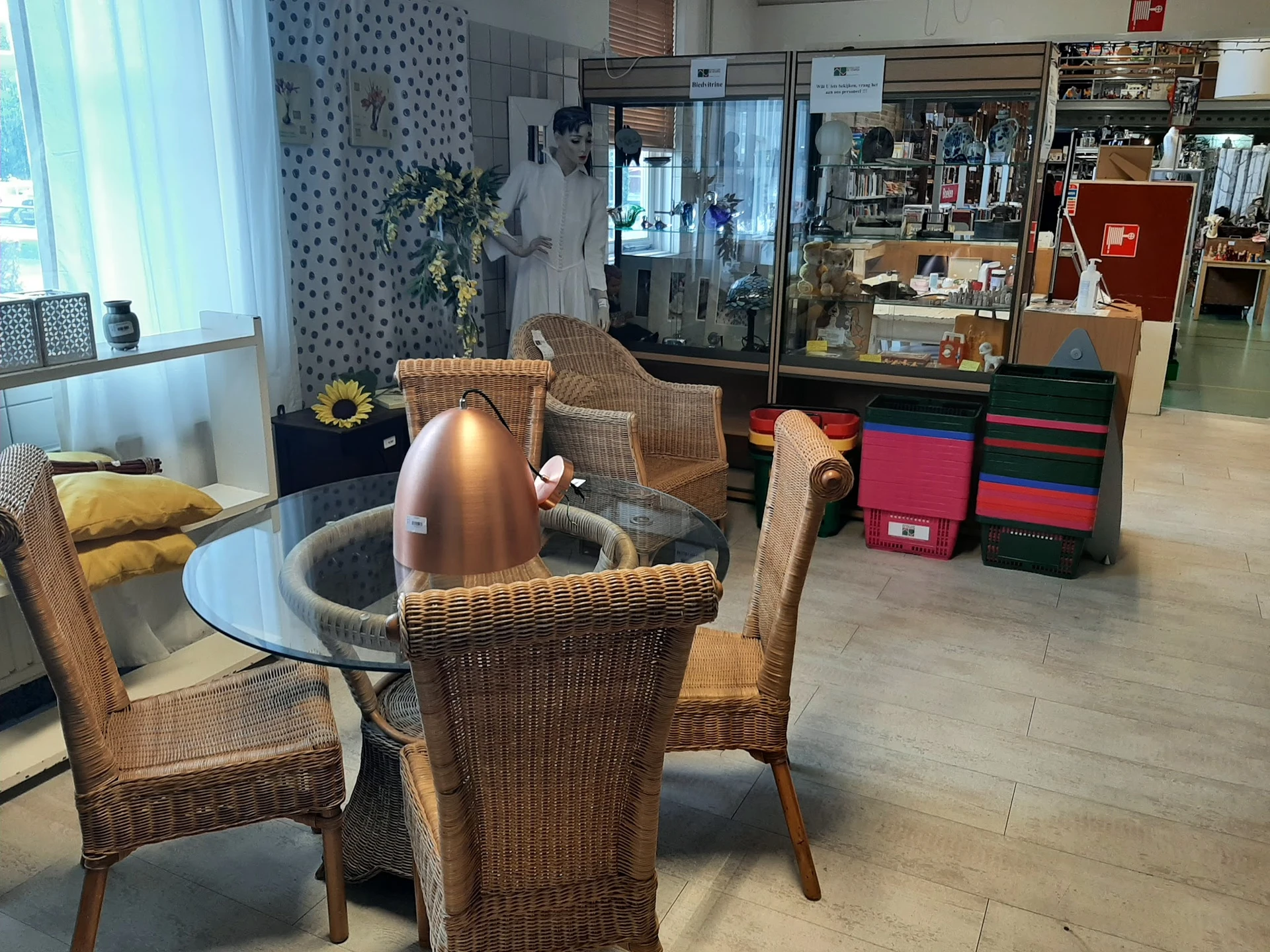 Zithoek met rieten stoelen rond glazen tafel in kringloopwinkel met stellingen op achtergrond