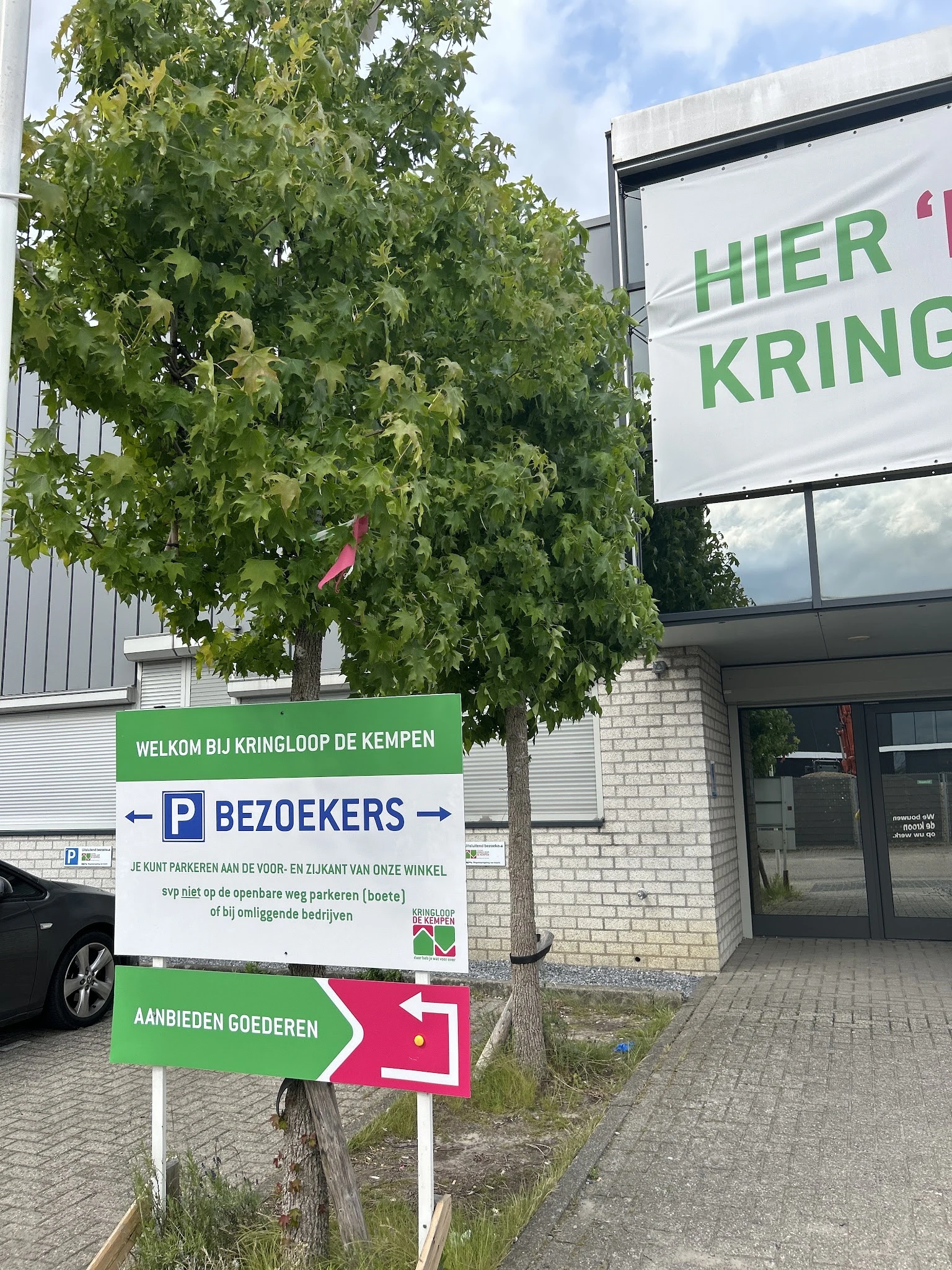 Ingang van kringloopwinkel met groen welkomsbord en parkeerplaatsen aanduiding