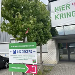 Ingang van kringloopwinkel met groen welkomsbord en parkeerplaatsen aanduiding