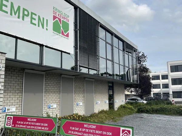 Voorgevel van kringloopwinkel met groene letters 'EMPEN!' en logo, witte gesloten deuren met parkeerplekken ervoor