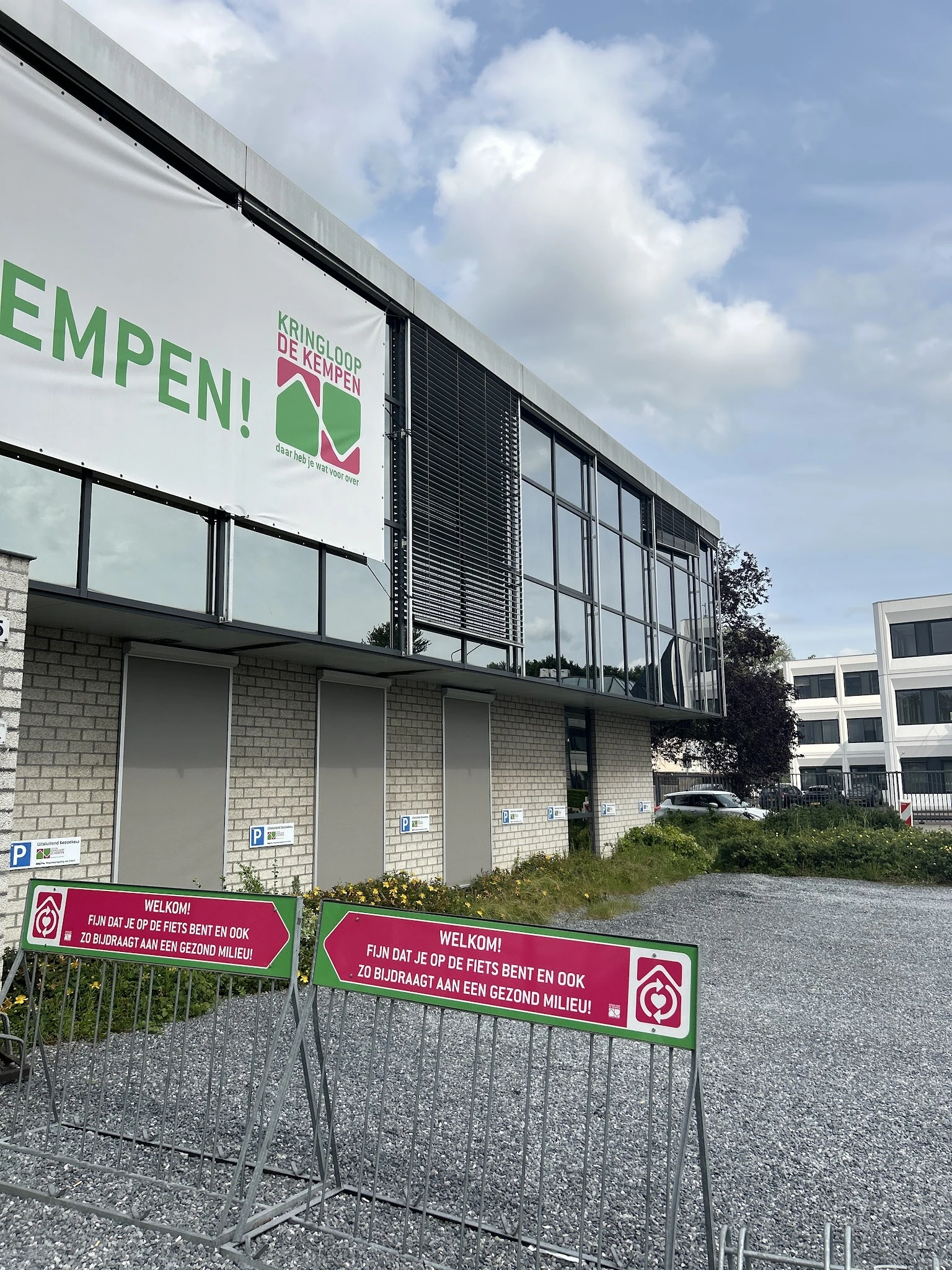 Voorgevel van kringloopwinkel met groene letters 'EMPEN!' en logo, witte gesloten deuren met parkeerplekken ervoor