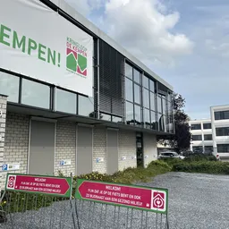 Voorgevel van kringloopwinkel met groene letters 'EMPEN!' en logo, witte gesloten deuren met parkeerplekken ervoor