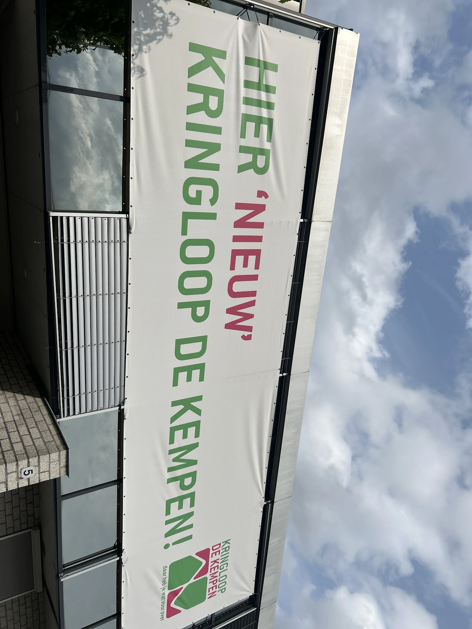 Zijkant van winkelgevel met wit doek waarop groene en rode tekst 'Kringloop De Kemp' zichtbaar