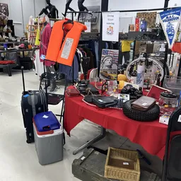 Rode toonbanktafel met tweedehands artikelen zoals schoenen, accessoires en huishoudspullen, met kleding en fietsen op de ach