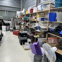 Binnenruimte met houten stellingen vol dozen, manden en diverse spullen; rommelig opgeslagen koopwaar