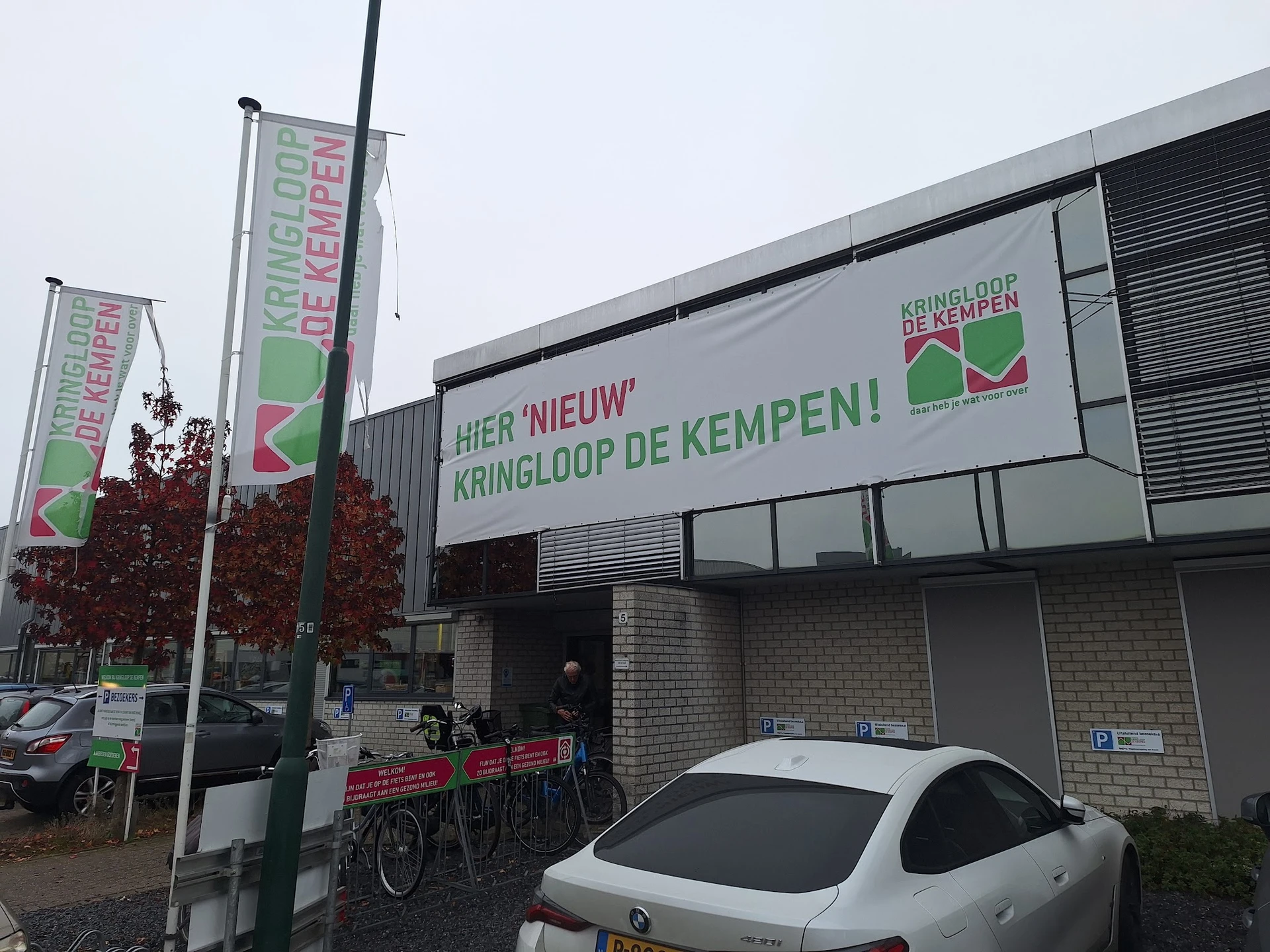 Voorgevel van kringloopwinkel met groene letters en logo, reclameborden en parkeerplaats