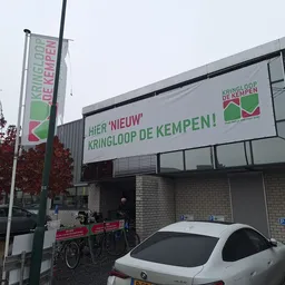 Voorgevel van kringloopwinkel met groene letters en logo, reclameborden en parkeerplaats