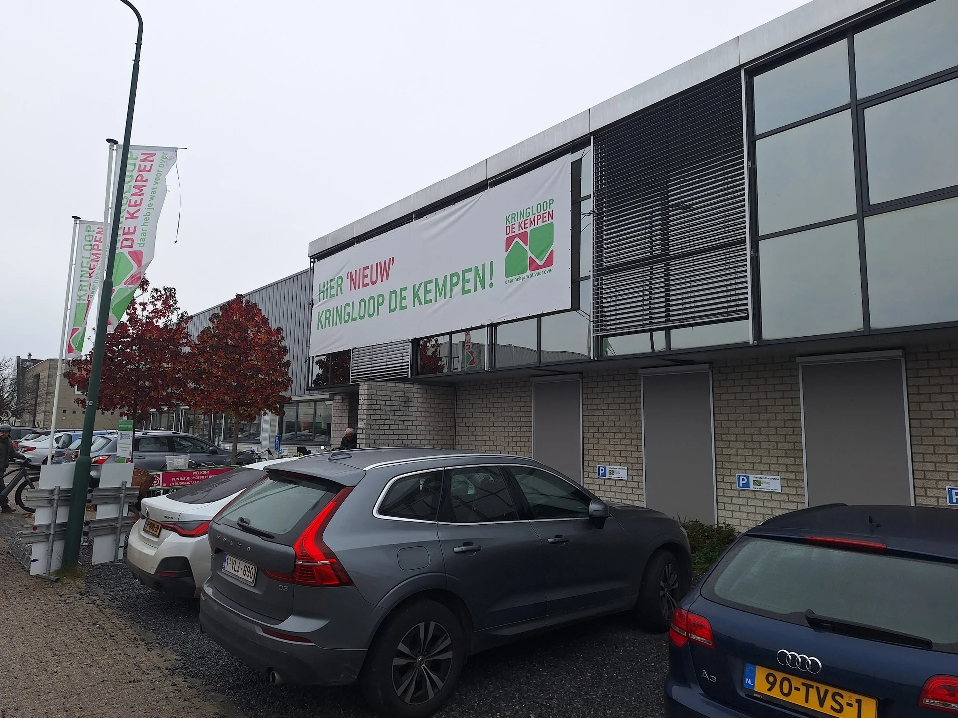 Gevel van kringloopwinkel met witte banner en grijze ramen, geparkeerde auto's op voorplein