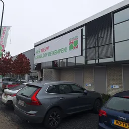 Gevel van kringloopwinkel met witte banner en grijze ramen, geparkeerde auto's op voorplein