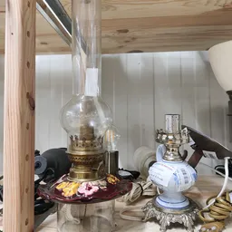 Vintage olielamp met messing fitting en glazen kap op een plank met andere huishoudelijke spullen