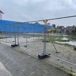 Bouwterrein met hekwerk, blauwe afdekking en bouwkraan achter metalen afzetting