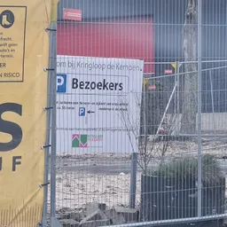 Ingang van kringloopwinkel met parkeerplaatsen-bord, gedeeltelijk verborgen door hekwerk en obstakels