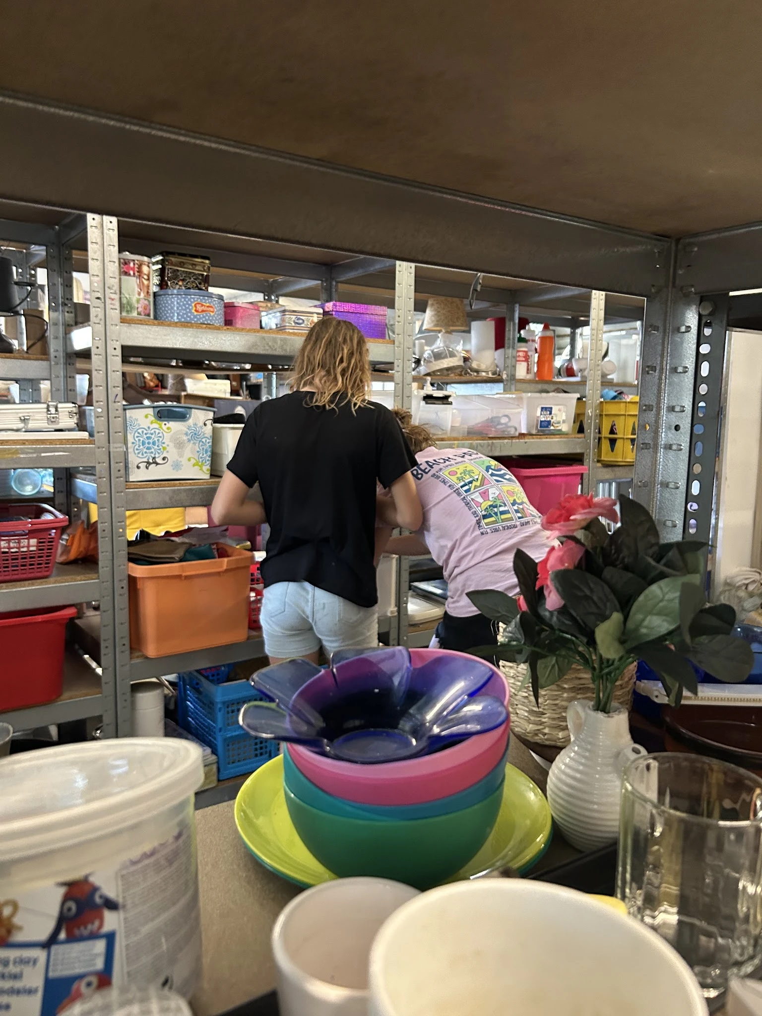 Binnenruimte van kringloopwinkel met stellingen, opbergboxen en diverse spullen; persoon bezig met inruimen