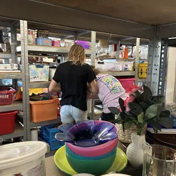 Binnenruimte van kringloopwinkel met stellingen, opbergboxen en diverse spullen; persoon bezig met inruimen