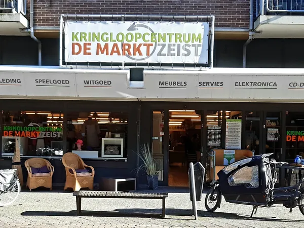 Gevel van kringloopwinkel De Markt Zeist met etalage, zitbanken en fietsen op het trottoir