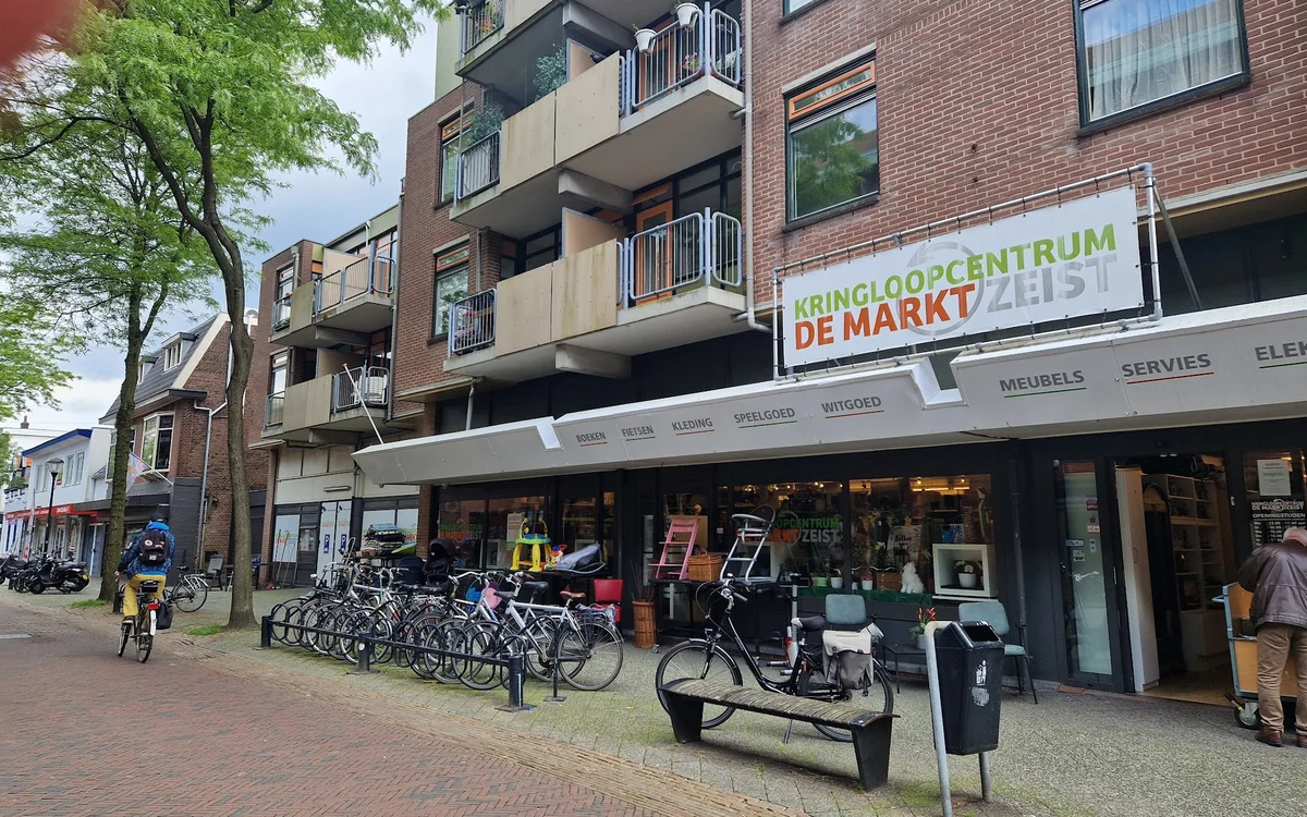 Gevel van kringloopwinkel met rode bakstenen, vitrines, fietsen voor de deur en naam 'DE MARKT' zichtbaar