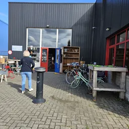 Exterieur van kringloopwinkel met donkergrijze gevel, rode en blauwe deuren, grote ramen, fietsenstalling en terrein met stoe