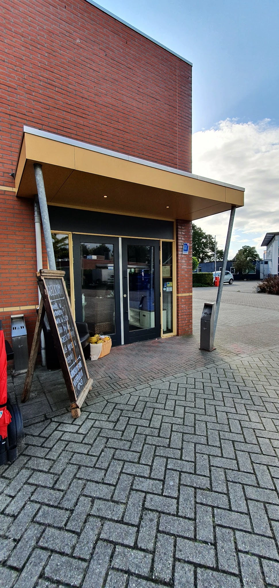Voorkant van kringloopwinkel met glazen deuren, rode bakstenen gevel en informatieborden op straat