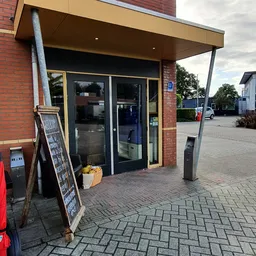 Voorkant van kringloopwinkel met glazen deuren, rode bakstenen gevel en informatieborden op straat