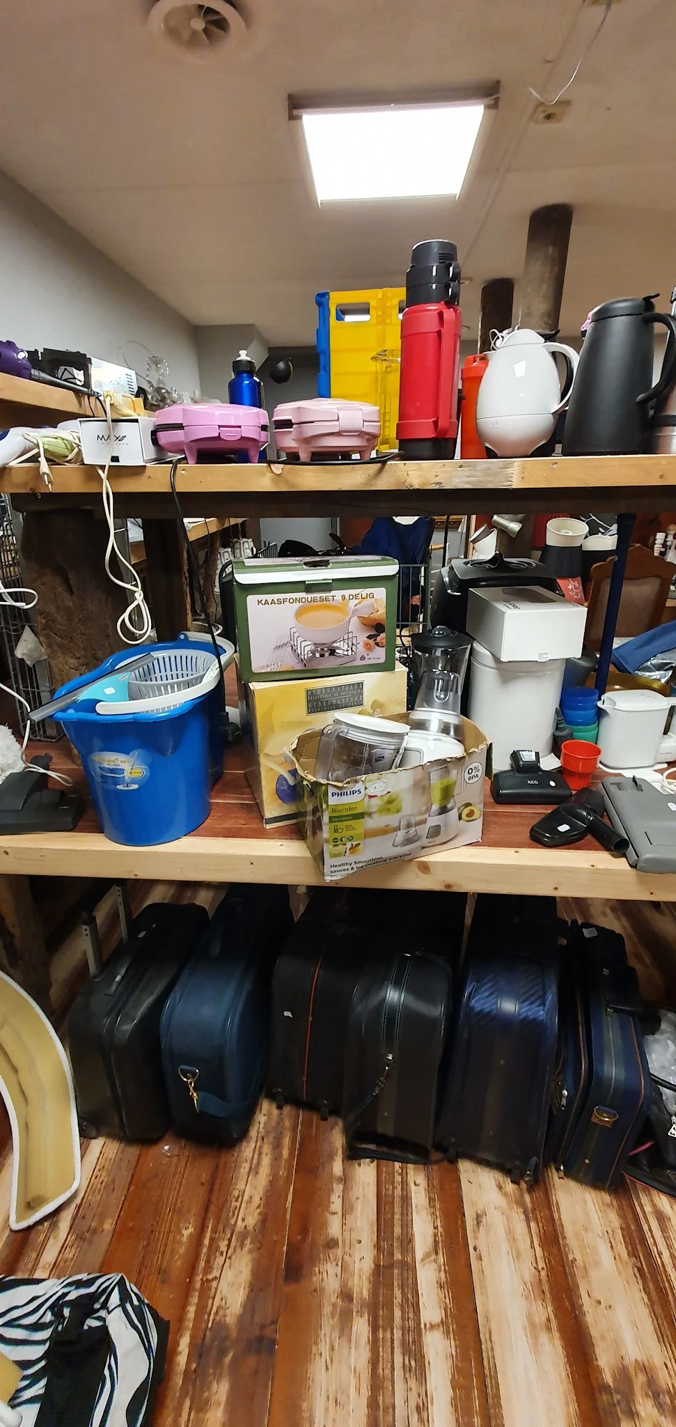 Diverse huishoudelijke apparaten en spullen op een houten plank, waaronder een blauwe emmer, een blender en andere keukenarti