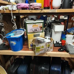 Diverse huishoudelijke apparaten en spullen op een houten plank, waaronder een blauwe emmer, een blender en andere keukenarti