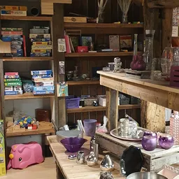 Houten stellingkast met diverse tweedehands huisartikelen: bordspellen, boeken, paarse serviesgoed, kristallen ornamentjes en