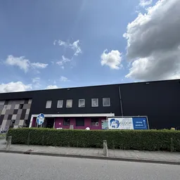 Voorkant van een moderne kringloopwinkel met zwart dak, grijze gevel en blauwe signage, omgeven door struiken