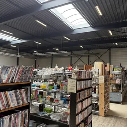 Magazijnhal met stellingen vol dvd's, cd's en boeken; voorgrond toont diverse kringloopwaar onder halogeenverlichting