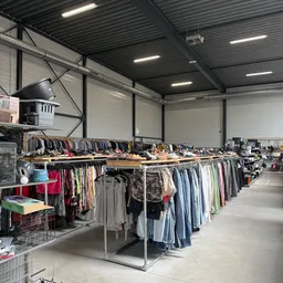 Magazijnruimte met lange rekken vol tweedehands kleding, hangers met jassen en shirts, en bakken met textiel op de grond