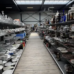 Binnenzicht van kringloopwinkel met lange doorgang tussen stellingen vol servies, glazenwerk en huishoudelijke artikelen