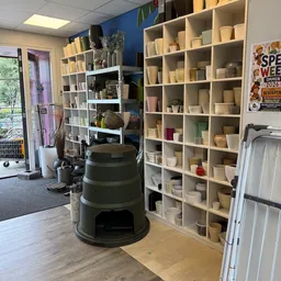Winkelinterieur met witte stellingen vol aardewerk, een groene compostwagen in het midden en een deur links