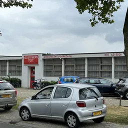 Gevel van kringloopwinkel met parkeerplaats en auto's ervoor, onder bewolkte hemel