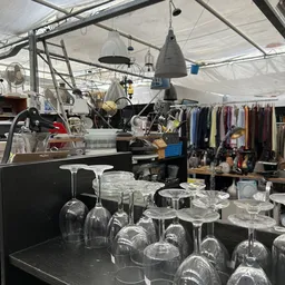 Uitgestalde glaswerk en serviesgoed op tafels in de kringloopwinkel, met kledingrekken op de achtergrond