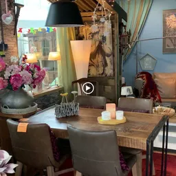 Woonkamer-achtige inrichting met houten tafel, grijze stoelen, bloemen, kasten en decoratieve objecten