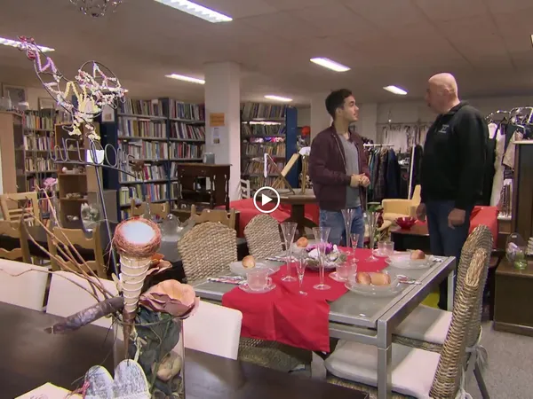 Winkelbinnenzicht met boekenkasten, kledingrekken en een tafel met roze tafelloper waarop twee personen zich bevinden
