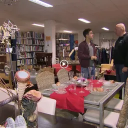 Winkelbinnenzicht met boekenkasten, kledingrekken en een tafel met roze tafelloper waarop twee personen zich bevinden